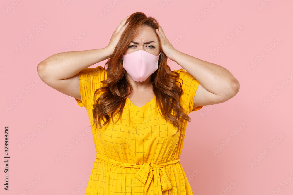Worried young redhead plus size body positive female woman in yellow dress face mask to safe from coronavirus virus covid-19 2019-ncov put hands on head isolated on pink background studio portrait.