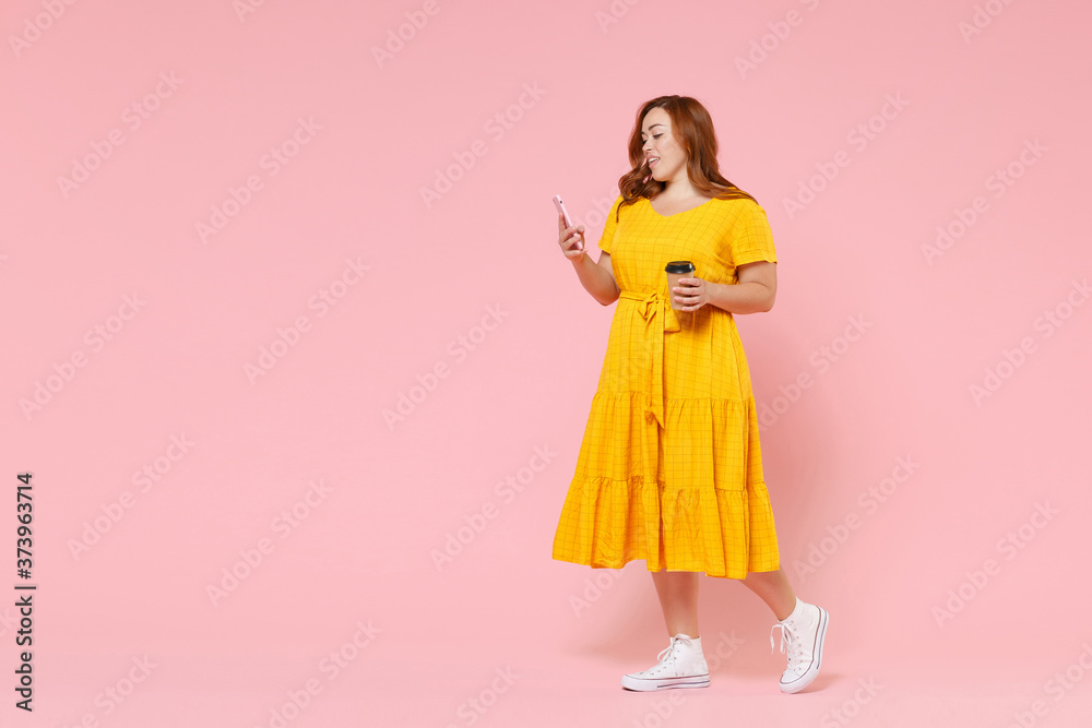 Full length portrait attractive young redhead plus size body positive female woman 20s in yellow dress hold paper cup coffee or tea using mobile phone isolated on pastel pink color background studio. Full length portrait attractive young redhead plus size body positive female woman 20s in yellow dress hold paper cup coffee or tea using mobile phone isolated on pastel pink color background studio.