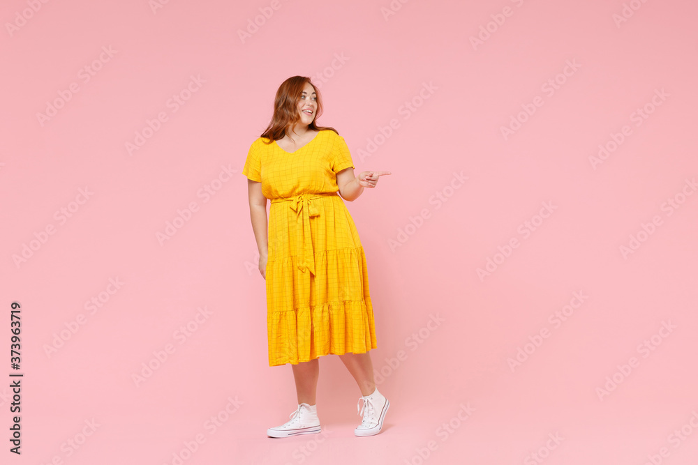 Full length portrait smiling young redhead plus size body positive female woman 20s in yellow dress pointing index finger aside on mock up copy space isolated on pastel pink color background studio. Full length portrait smiling young redhead plus size body positive female woman 20s in yellow dress pointing index finger aside on mock up copy space isolated on pastel pink color background studio.