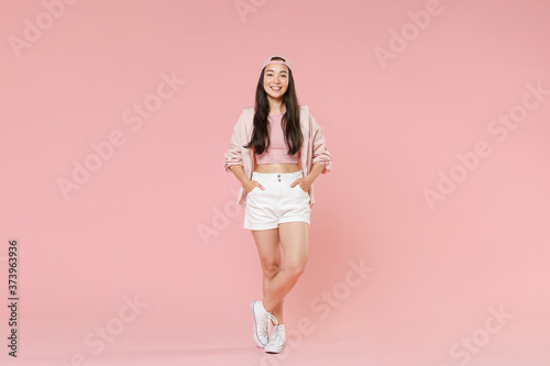 Full length portrait of smiling young asian girl in casual clothes, cap isolated on pastel pink wall background studio portrait. People lifestyle concept. Mock up copy space. Holding hands in pockets.