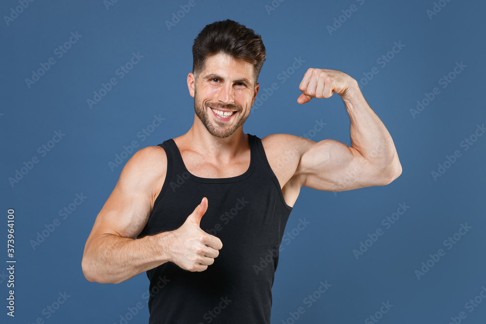 Smiling young bearded fitness sporty guy sportsman in black undershirt isolated on blue wall background studio portrait. Workout sport motivation lifestyle concept. Showing biceps muscles thumb up. Smiling young bearded fitness sporty guy sportsman in black undershirt isolated on blue wall background studio portrait. Workout sport motivation lifestyle concept. Showing biceps muscles thumb up.