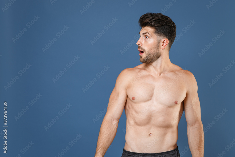 Fototapeta premium Shocked young bearded fitness sporty strong guy bare-chested muscular sportsman posing isolated on blue background studio. Workout sport motivation lifestyle concept. Keeping mouth open looking aside.