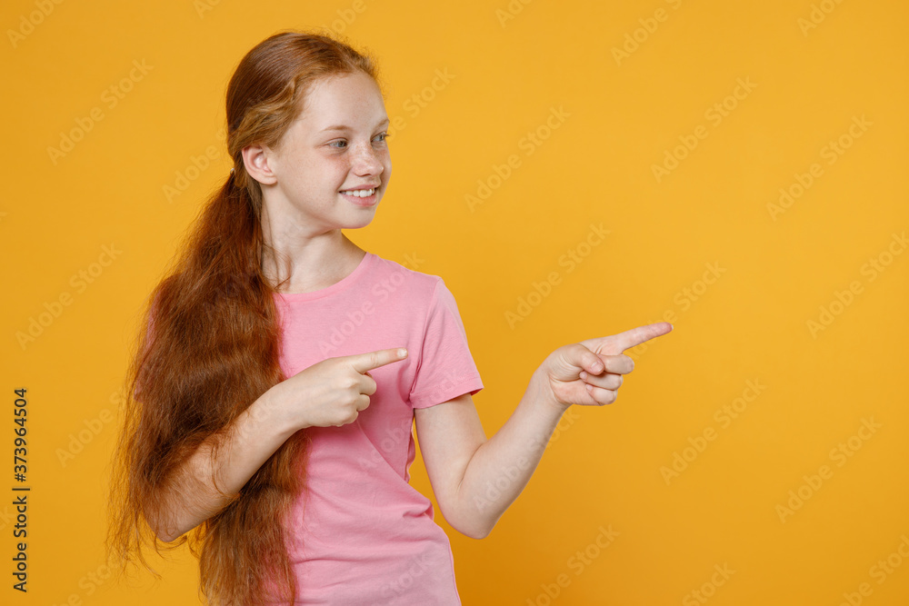 Smiling little ginger redhead kid girl 12-13 years old in pink casual t-shirt posing pointing index fingers aside on mock up copy space isolated on yellow color background children studio portrait. Smiling little ginger redhead kid girl 12-13 years old in pink casual t-shirt posing pointing index fingers aside on mock up copy space isolated on yellow color background children studio portrait.