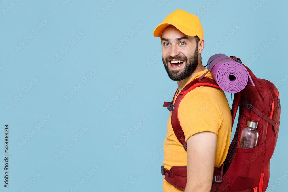 Side view excited traveler young man in yellow casual t-shirt cap with backpack isolated on blue background. Tourist traveling on weekend getaway. Tourism discovering hiking concept. Looking camera. Side view excited traveler young man in yellow casual t-shirt cap with backpack isolated on blue background. Tourist traveling on weekend getaway. Tourism discovering hiking concept. Looking camera.