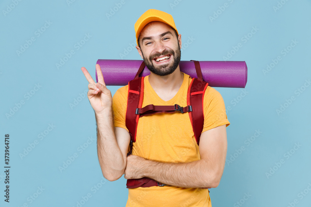 Smiling traveler young man in yellow casual t-shirt cap with backpack isolated on blue wall background. Tourist traveling on weekend getaway. Tourism discovering hiking concept. Showing victory sign. Smiling traveler young man in yellow casual t-shirt cap with backpack isolated on blue wall background. Tourist traveling on weekend getaway. Tourism discovering hiking concept. Showing victory sign.