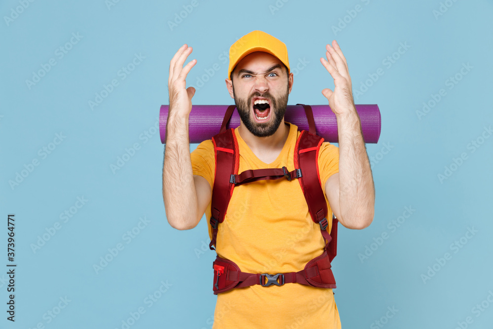 Angry traveler young man in yellow t-shirt cap with backpack isolated on blue background. Tourist traveling on weekend getaway. Tourism discovering hiking concept. Spreading hands screaming swearing.