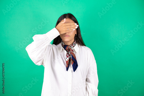 Young beautiful business woman over isolated green background serious and covering her eyes with her hand