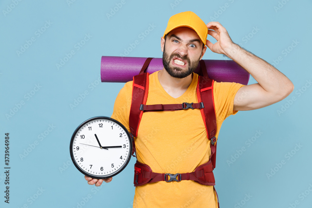 Preoccupied young traveler man in yellow casual t-shirt cap backpack isolated on blue background. Tourist traveling on weekend getaway. Tourism discovering hiking concept. Hold clock put hand on head.