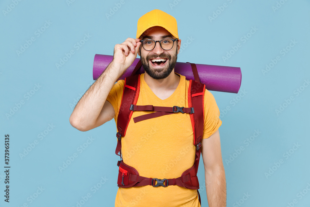 Cheerful traveler young man in yellow casual t-shirt cap glasses with backpack isolated on blue background. Tourist traveling on weekend getaway. Tourism discovering hiking concept. Looking camera. Cheerful traveler young man in yellow casual t-shirt cap glasses with backpack isolated on blue background. Tourist traveling on weekend getaway. Tourism discovering hiking concept. Looking camera.