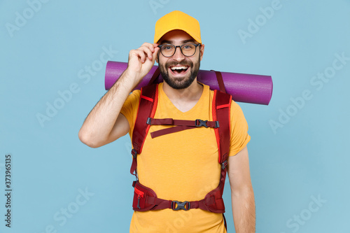 Cheerful traveler young man in yellow casual t-shirt cap glasses with backpack isolated on blue background. Tourist traveling on weekend getaway. Tourism discovering hiking concept. Looking camera.