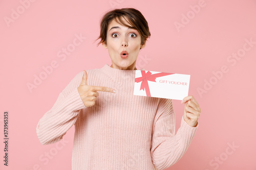 Shocked young brunette woman girl in knitted casual sweater posing isolated on pastel pink background studio portrait. People emotions lifestyle concept. Pointing index finger on gift certificate.