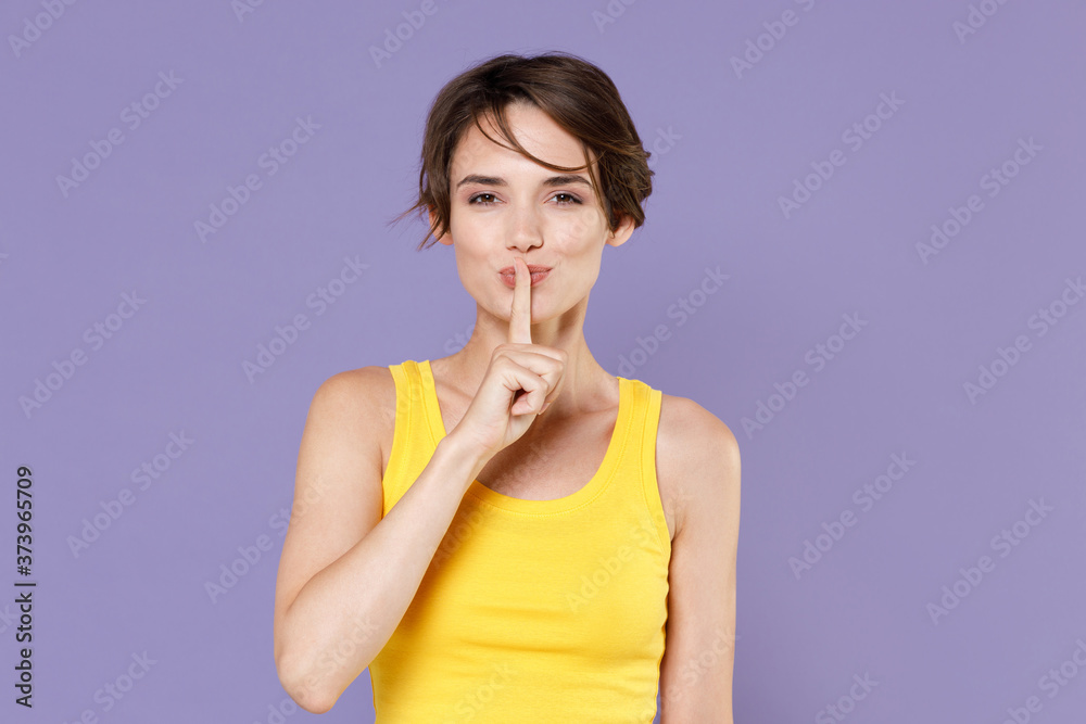 Secret young brunette woman girl in yellow casual tank top posing isolated on pastel violet background studio portrait. People lifestyle concept. Saying hush be quiet with finger on lips shhh gesture. Secret young brunette woman girl in yellow casual tank top posing isolated on pastel violet background studio portrait. People lifestyle concept. Saying hush be quiet with finger on lips shhh gesture.