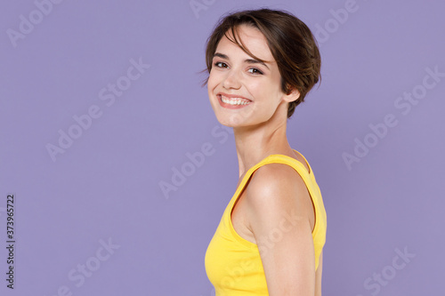 Side view of smiling young brunette woman girl wearing yellow casual tank top posing isolated on pastel violet background studio portrait. People sincere emotions lifestyle concept. Looking camera.