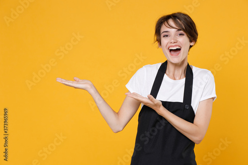 Foto Cheerful young female woman 20s barista bartender barman employee in white casual t-shirt apron pointing hands aside on mock up copy space isolated on yellow color wall background studio portrait