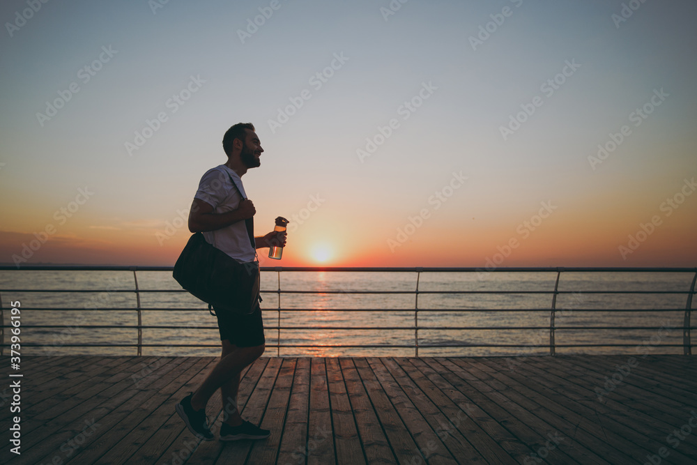 Full length portrait of smiling young bearded athletic man guy 20s in white t-shirt with sports bag posing coming to training hold bottle of water looking aside at sunrise over the sea outdoors.