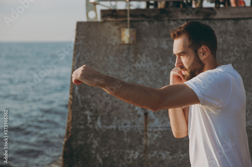 Portrait side view of handsome attractive young athletic boxer man guy 20s in casual white t-shirt black shorts posing training doing boxing exercises looking aside at sunrise over the sea outdoors.