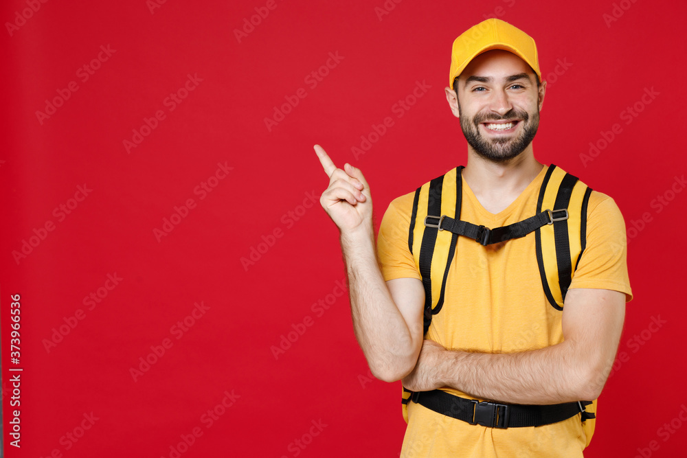 Delivery man 20s in yellow cap t shirt uniform thermal bag backpack points finger aside isolated on red background studio Guy male employee work as courier Service coronavirus covid-19 virus concept Delivery man 20s in yellow cap t shirt uniform thermal bag backpack points finger aside isolated on red background studio Guy male employee work as courier Service coronavirus covid-19 virus concept