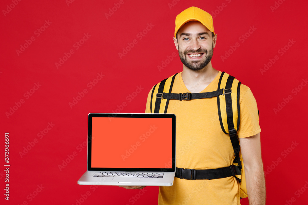 Delivery man in yellow cap t-shirt uniform thermal bag backpack with food hold laptop pc isolated on red background studio Guy male employee work as courier Service coronavirus covid-19 virus concept