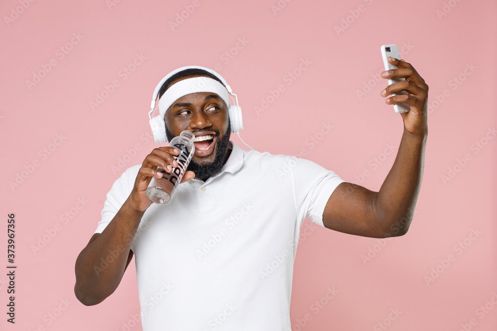 Φωτογραφία Funny young african american fitness sports man 20s in headband t-shirt listen music with headphones doing selfie shot on mobile phone hold bottle of water isolated on pink background studio portrait