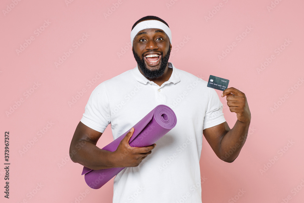 Φωτογραφία Excited young bearded african american fitness sports man in white headband t-shirt posing hold yoga mat credit bank card spending time in gym isolated on pastel pink color background studio portrait