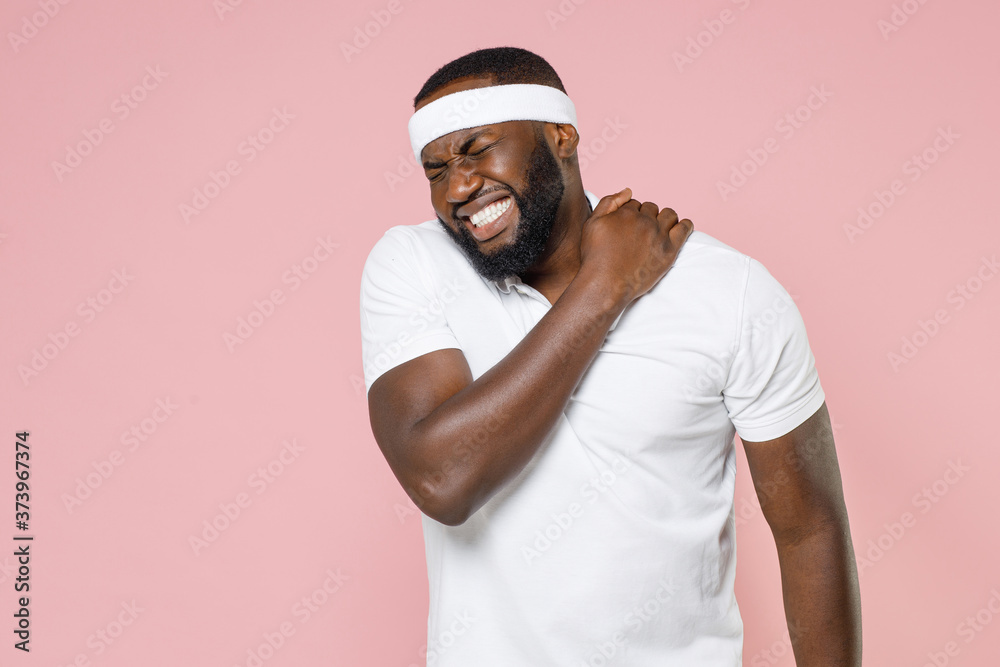 Sick injured young bearded african american fitness sports man in headband t-shirt feels bad pain spasm back sprain or neck spending time in gym isolated on pink color wall background studio portrait. Sick injured young bearded african american fitness sports man in headband t-shirt feels bad pain spasm back sprain or neck spending time in gym isolated on pink color wall background studio portrait.