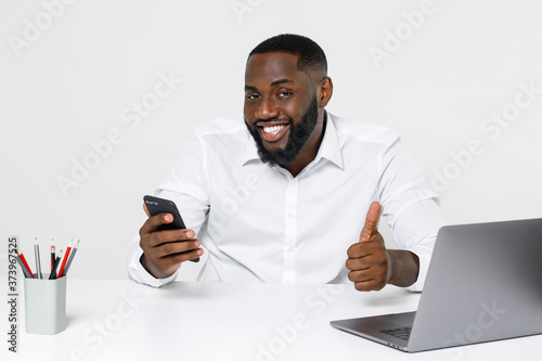 Smiling young african american male business man in white shirt posing working in office sitting at desk with laptop pc using mobile cell phone showing thumb up isolated on white color background. © ViDi Studio