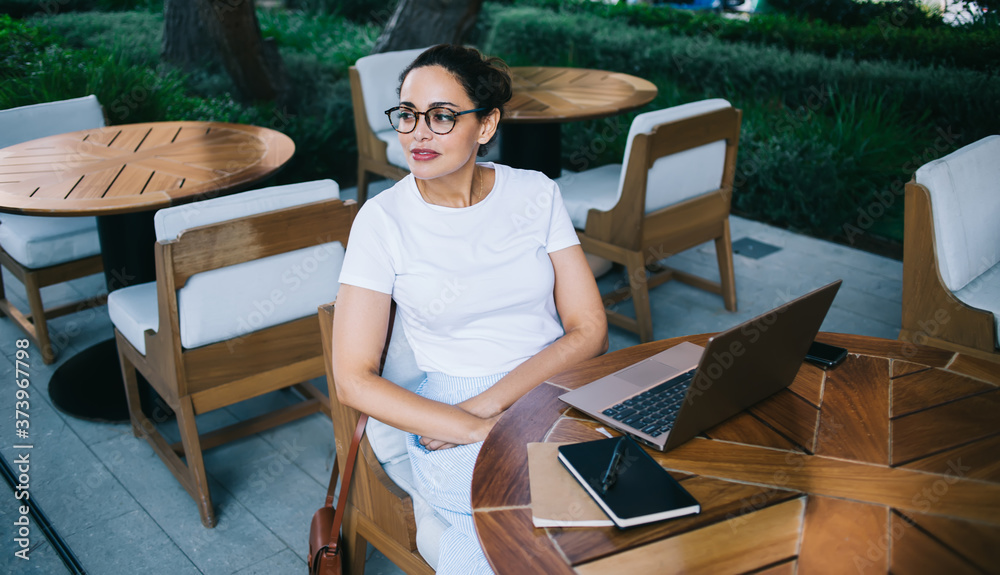Confident woman in eyeglasses sitting at table outside modern workspace with laptop and documents