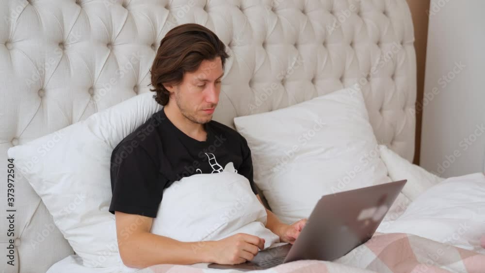 Young man with long hair lying in the bed and trying to work on the laptop, tired and nod off and nap. White linens. Black shirt one the guy. Bright bedroom