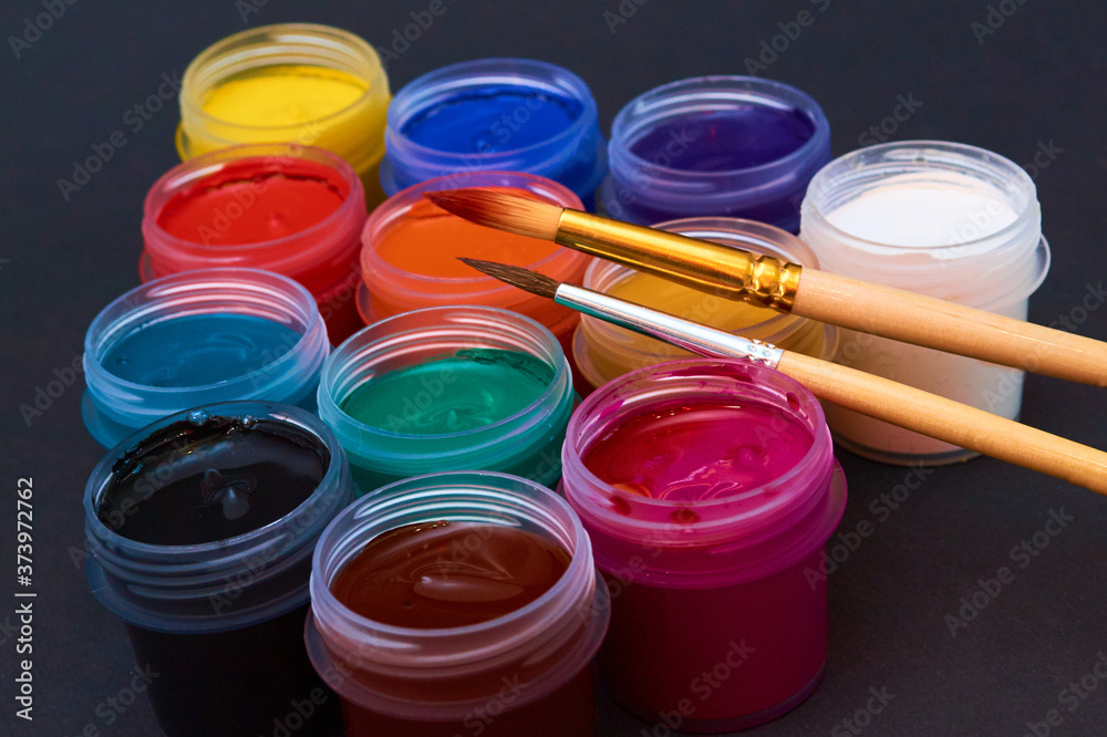 multicolored jars with paint gouache and a brush, on a black background close-up multicolored jars with paint gouache and a brush, on a black background close-up