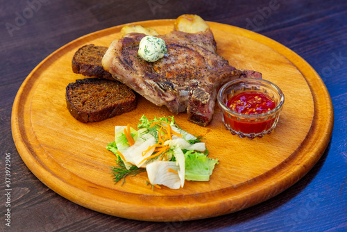 
freshly grilled Tomahawk steak on a round wooden plate with tomato sauce, slices of black bread, lettuce and carrots. Close view