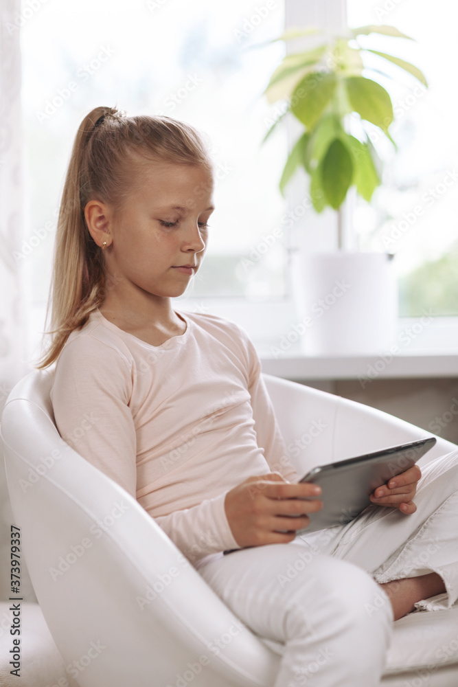 Beautiful teenage girl sitting in white armchair in white room doing homework using tablet, online education, distance learning, technology concept Beautiful teenage girl sitting in white armchair in white room doing homework using tablet, online education, distance learning, technology concept