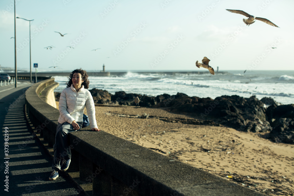 Young multi-cultural woman sitting of sea promenade. Seagulls fly over the foaming surf. Young multi-cultural woman sitting of sea promenade. Seagulls fly over the foaming surf.