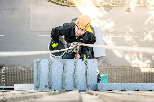 Male worker hanging on rope access and height construction project. Working on building wall insulation and facade painting decoration job.