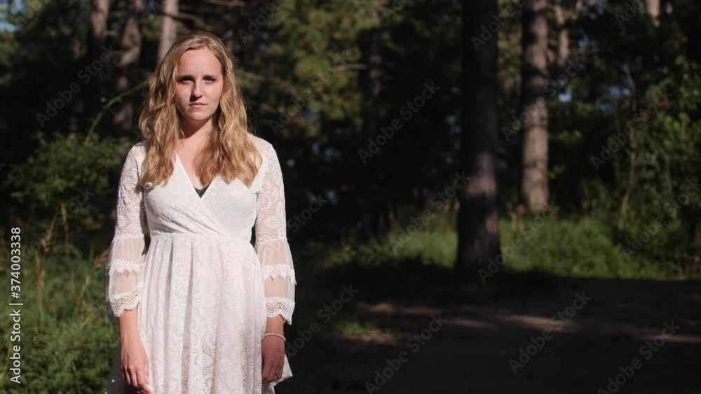 Beautiful young blond woman in white dress looks into camera in forest medium wide-shot Beautiful young blond woman in white dress looks into camera in forest medium wide-shot