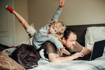 Work from home with kids children. Father working on laptop in bedroom with child daughter on his back. Funny candid family moments. New normal during coronavirus self-isolation quarantine.