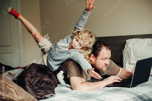 Work from home with kids children. Father working on laptop in bedroom with child daughter on his back. Funny candid family moments. New normal during coronavirus self-isolation quarantine.