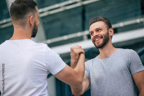 Men s friendly handshake. Two young bearded handsome friends in casual t-shirts are posing together on the city background.