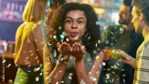 Attractive mixed race young celebrating woman blowing confetti to camera while posing in the night club. Friends chatting, having drinks at the bar counter in the background