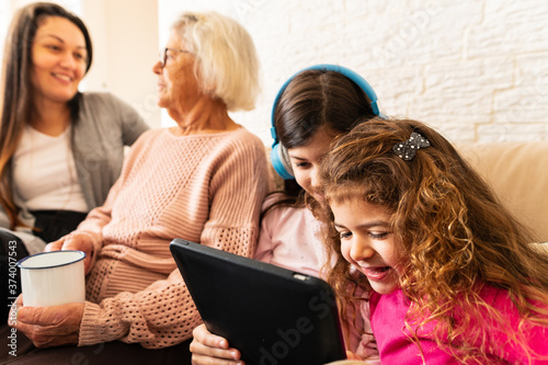 Lovely Young granddaughters with digital tablet having fun and mother and grandmother talking in the back Indoors at home living room. Relationship, leisure, enjoyment, aging concept.