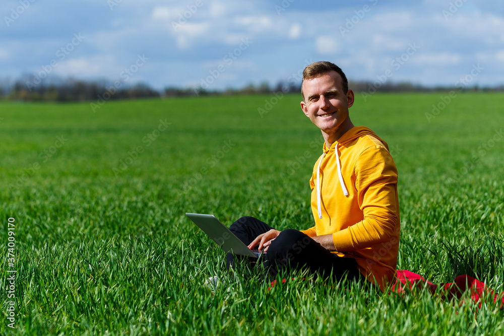 Young businessman sitting on green grass and using laptop computer. Handsome man working with computer in park at sunny summer day. Outdoors nature journey and relaxation. Freelance work concept Young businessman sitting on green grass and using laptop computer. Handsome man working with computer in park at sunny summer day. Outdoors nature journey and relaxation. Freelance work concept
