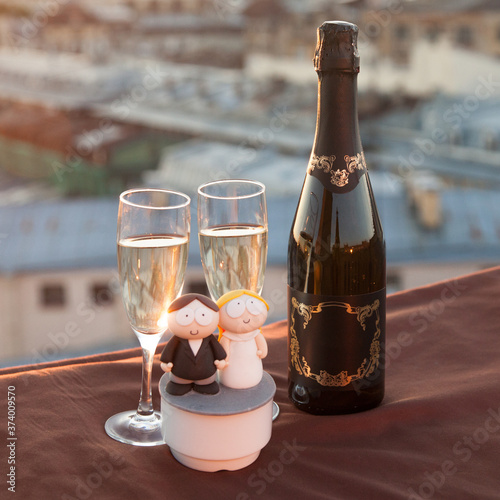 A bottle and two glasses of champagne along with wedding figurines against the backdrop of city rooftops at sunset