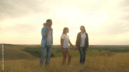 healthy smiling family, holding hands, walking across field at sunset in mountains, little daughter riding on daddy's back. happy children and parents walk in rays of beautiful sun, travel on vacation