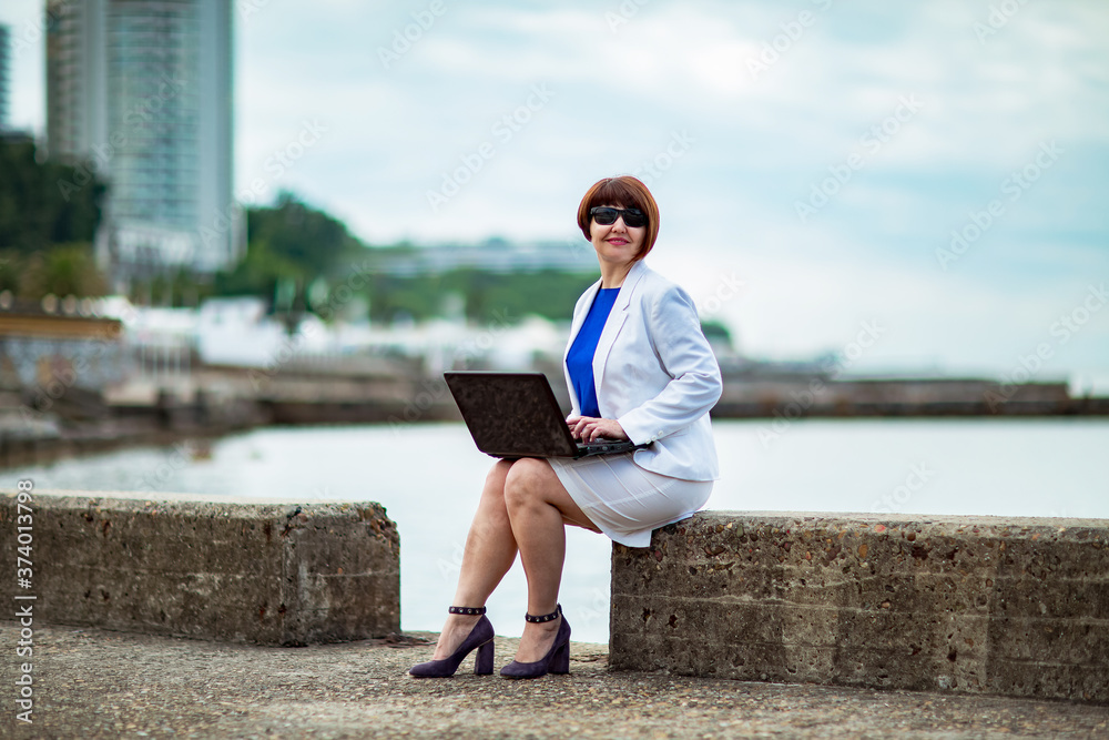 A beautiful woman, brown-haired, in a white business suit and blue blouse, working on a laptop against the background of the city and the sea