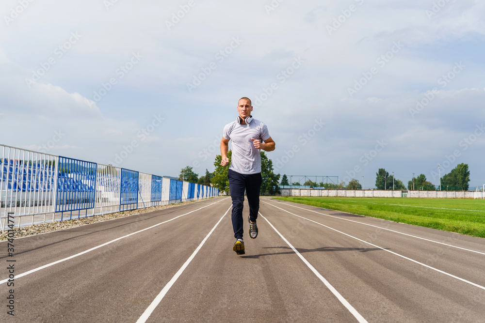 Full length front view on adult caucasian man jogging on the running track - Blonde male athlete in stadium training in summer day - real people healthy lifestyle concept