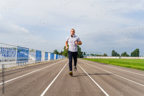 Full length front view on adult caucasian man jogging on the running track - Blonde male athlete in stadium training in summer day - real people healthy lifestyle concept