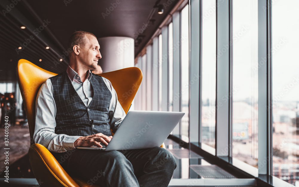 A handsome cheerful caucasian man entrepreneur is sitting with a laptop on his knees on an orange armchair next to the window indoors of a luxurious interior of a modern business office skyscraper - obrazy, fototapety, plakaty 