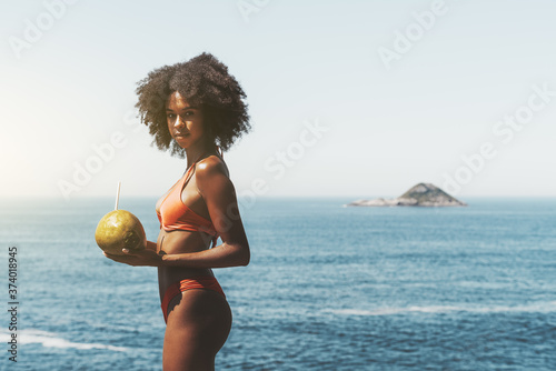 A portrait of a ravishing African-American girl standing in a swimsuit on the coast of a tropical resort with a coconut with a straw in her hand; an island and a copy space place on the right