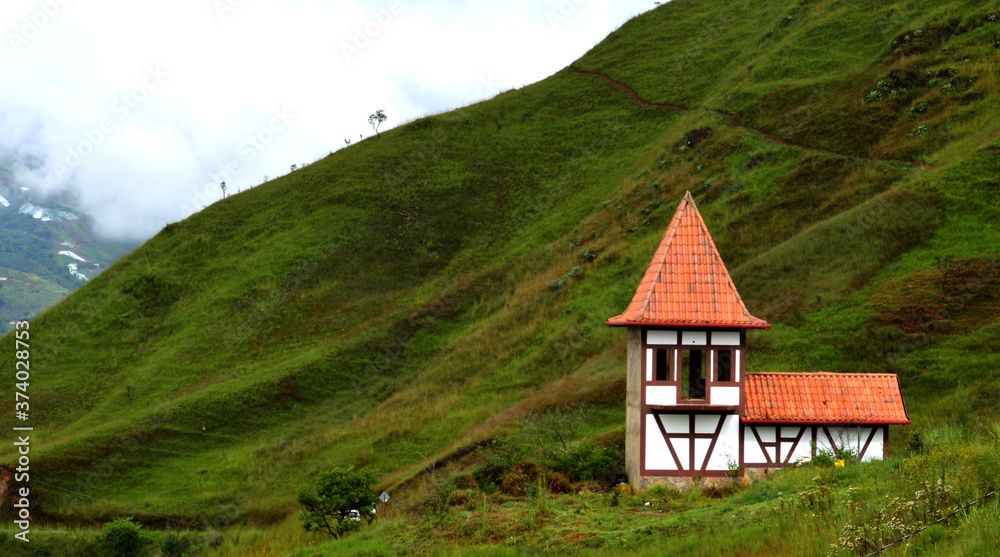 A small house with German architecture, open windows, white walls with brown stripes, and an orange roof. Located on the mountain of green grass, low trees, narrow trails and behind the cloudy sky.