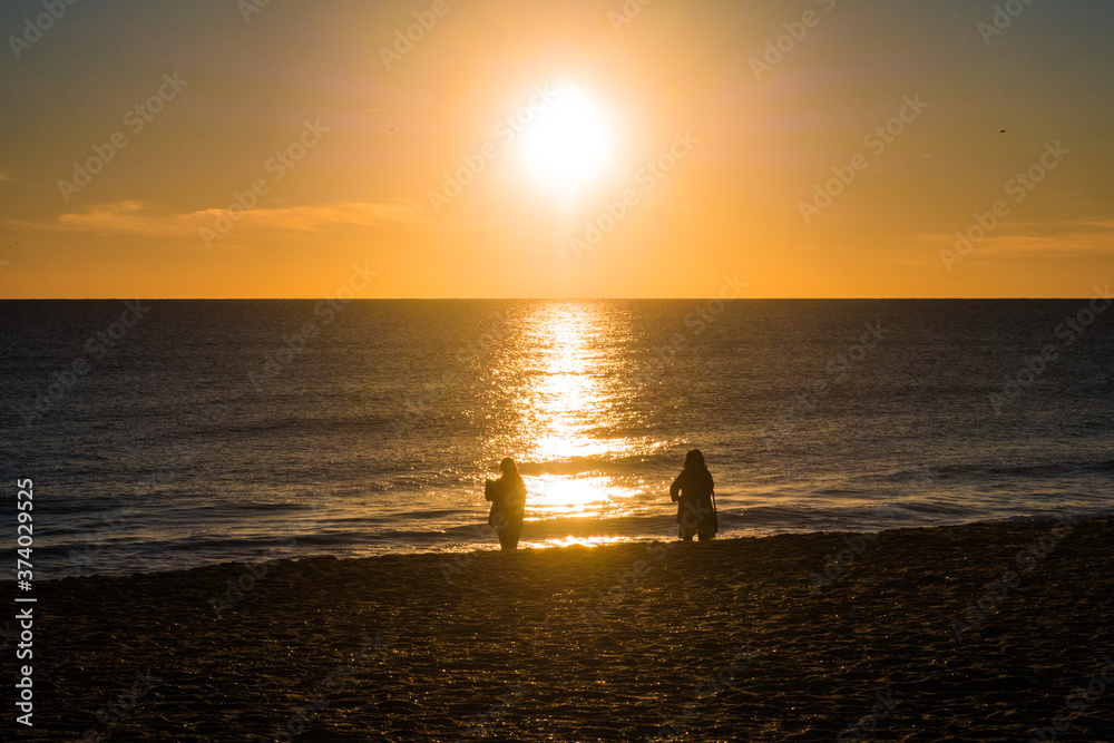Sunset at the beach with sun in the sky and 2 people in silhouette