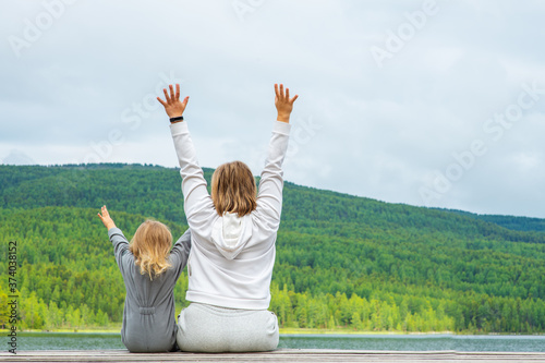 A woman and her child sit with their back to the camera with their hands raised against the backdrop of a mountain lake and mountains in the Altai Republic in Russia